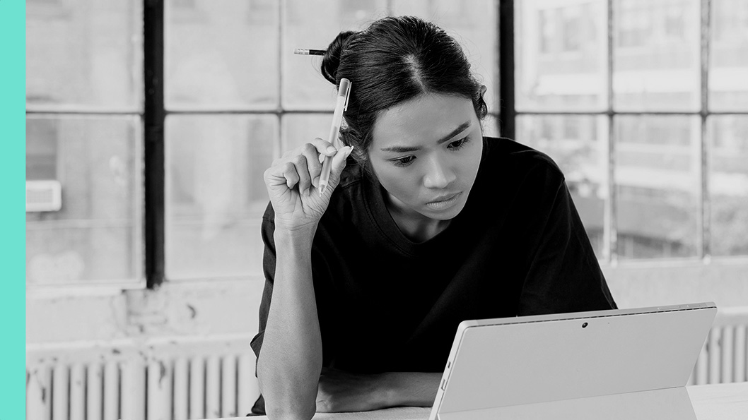 Individual seated in front of a laptop, resting a pen against their hair while looking at the screen.