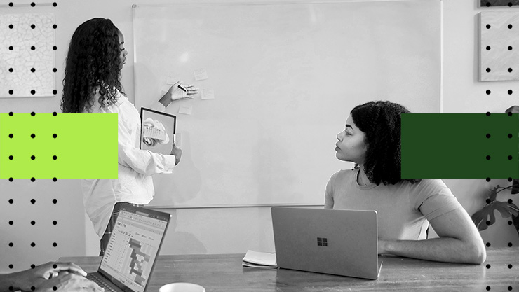 One person presents sticky notes on a whiteboard while another, seated at a laptop, follows along.