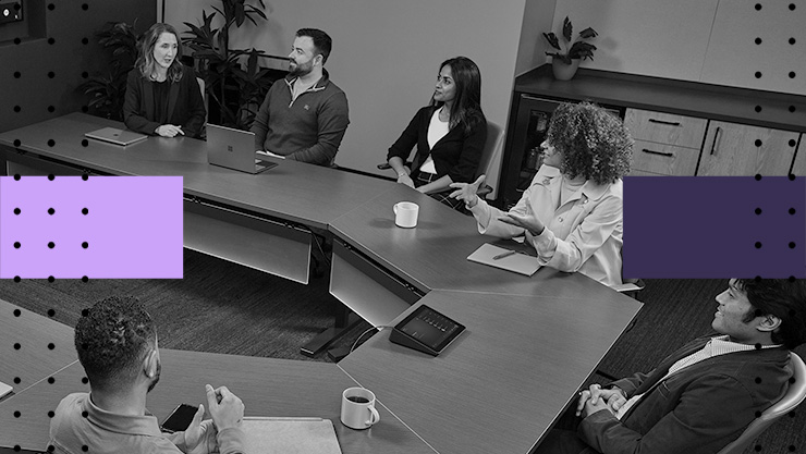 A group of people sit around a conference table in a modern meeting room while viewing remote participants on a large video screen.